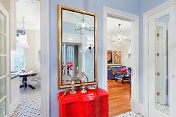 Red-painted, postmodern chest of drawers below gilt-framed mirror on wall painted pale blue flanked by open interior doors with view of rooms beyond