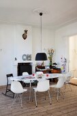 Black pendant lamp above white, oval dining table and shell chairs; fireplace and workspace in background