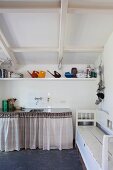 Giesskannen und Küchenutensilien auf Bord unter einer Dachschräge, darunter der Spülenschrank mit Vorhang und seitlich eine alte Truhenbank
