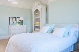 French bed with scatter cushions and white bedspread and white-painted, carved, rustic linen cupboard in pastel bedroom in period apartment