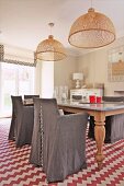 Chairs with grey loose covers around dining table below wicker pendant lamps and rug with red and white zigzag patterns in country-house interior