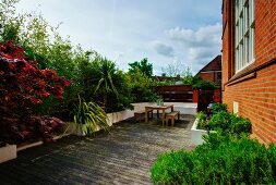 Bushes and palm trees in raised beds surrounding wooden terrace with table and benches adjoining brick facade of converted church