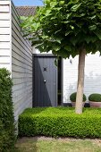 Anthrazitfarbene Schiebetür und holzverschalter Anbau im Vorgarten eines Ferienhausses mit Baum inmitten niedriger Buchshecke