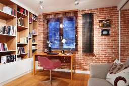 Simple workspace below window; pink swivel chair and wooden desk against brick wall, wooden, fitted shelving with painted fronts to one side