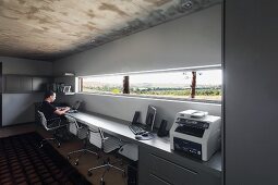 weiße Büro Drehstühle vor langer, zwischen den Wänden eingespannter Arbeitstisch, an Wand mit schmalem Fensterband und Landschaftsblick