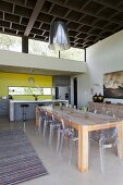Rustic, solid wood table and Ghost chairs; open-plan kitchen with yellow wall in background and coffered-style ceiling