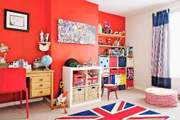 Kinderzimmer mit roter Wand und Teppich mit Union-Jack-Flagge, seitlich Schreibtisch aus Holz, in Nische Regaleinbau mit farbigen Aufbewahrungsboxen
