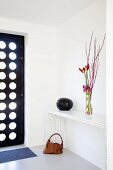 Vase of amaryllis and branches on modern, white console table next to black, modern front door with many bullseye windows