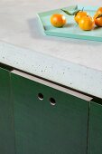 Close-up of DIY kitchen counter with concrete worksurface and hole pulls in dark green doors; tangerines on tray