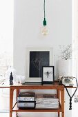 Minimalist, vintage pendant lamp above vintage telephone next to framed pictures on fifties-style console table against narrow section of wall