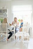 Mother, daughter and baby in high chair at dining table in white, country-house kitchen