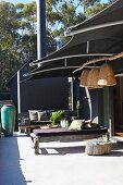 Rustic wooden benches with seat cushions on modern terrace below dark fabric awning with straw lampshades