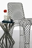 Stuhl aus Maschendrahtgewebe, Beistelltisch mit spiralförmig gebogenen Fussstreben und Gerberablüte in korallenartig konstruierter Vase