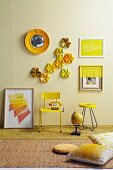 Still-life arrangement of small, yellow table and yellow-painted, retro chair below cake tins and pictures on pastel yellow wall