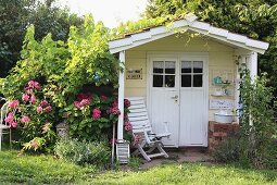 Romantisch bewachsenes Holzgartenhäuschen pastellgelb gestrichen, unter Vordach weißer Armlehnstuhl
