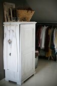 White dress on coat hanger hung on white-painted wardrobe with basket on top; clothes rail in background