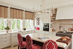 weiße Landhausküche mit freistehender Theke, gepolsterten Barhockern mit rotweißem Brokatmuster, darüber weisser Kronleuchter