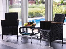 Rattan chairs flanking fruit bowl on retro side table in front of floor-to-ceiling windows with view of pool in garden