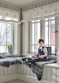 Little boy sitting on sheepskin blanket on window seat in wood-clad conservatory
