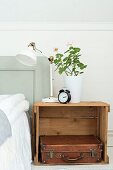Retro table lamp and geranium in white pot on wooden crate used as bedside table holding leather suitcase