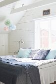 Scatter cushions and blankets on bed below window in wood-clad attic room decorated with pastel paper pompoms