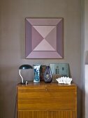 Table lamp and blue and white vase on bureau below geometric artwork on mauve wall
