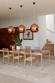 Long designer dining table and chairs made from pale wood under copper-coloured lampshades in modern dining area