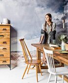 Young woman next to rustic wooden table, wooden chair, white-painted chair and chest of drawers in front of mural of grey cloudy sky