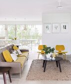 Loungebereich mit Sofa und Klassikerstuhl gelb lackiert um Couchtisch in offenem Wohnraum, im Hintergrund Essplatz vor Fensterfront