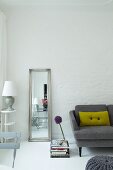 Full-length mirror leaning against whitewashed brick wall between lamp on side table and grey sofa