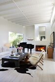 Chairs with sheepskin blankets and white sofa around coffee table; grand piano next to open fireplace in background in living room