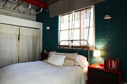 Bed with vintage metal frame below window and table lamp on red bedside cabinet against brick wall painted dark green