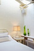 Lamp on bedside table between bed and green vases on shelf in corner of room with whitewashed brick walls