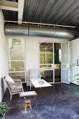 Terrace furnished with wicker chair, stool and small table outside contemporary house with ventilation duct running below corrugated metal ceiling
