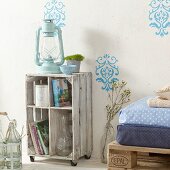 Upcycled cabinet made from wine crate on castors with hurricane lamp on top against wall with light blue stencilled pattern