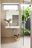 Sink on bathroom wall covered in round, beige iridescent mosaic tiles; walk-in shower with glass wall looking into planted courtyard