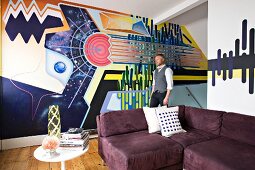 Artist in front of creative, graphic mural; aubergine corner sofa and vintage side table