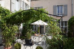Sonnenbeschienener Terrassenplatz unter idyllisch berankter Metallpergola im Innenhof eines französischen Landhauses