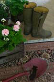 Soil on spade and wooden crate of flowering geraniums on rug next to Wellington boots