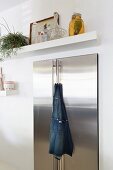 Cooks' apron hanging from handle of stainless steel fridge below white floating shelf
