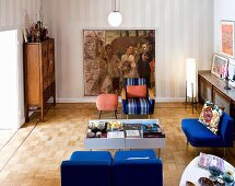 Blue retro easy chairs around display-case tables and painting on striped wallpaper in eclectic living room with mosaic parquet floor