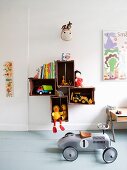 Retro, ride-on toy car on grey wooden floor and toys and books in and on wooden crates mounted on wall