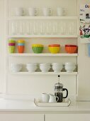 White and coloured crockery on white-painted, wall-mounted shelves above cafetiere on tray on worksurface