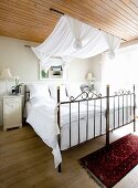 Double bed with metal frame, white bed linen and fabric canopy hanging from rods suspended from wooden ceiling