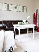 White-painted side table with turned legs in front of black leather couch below framed botanical pictures on wall