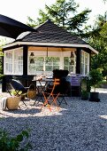 Wooden summerhouse painted black with white lattice windows behind colourful folding chairs on gravel terrace in