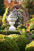 Antique, stone Greek urn in landscaped garden with low box hedges and old wrought iron gate in background