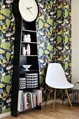 Long-case clock with shelves in black casing, tins with contrasting retro patterns, classic-style shell chair and leaf-patterned wallpaper