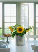 Sunflowers in metal container next to pastries on cake stand on white table