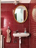 Waschbecken unter ovalem Spiegel mit Goldrahmen an roter Fliesenwand in moderner Badezimmerecke